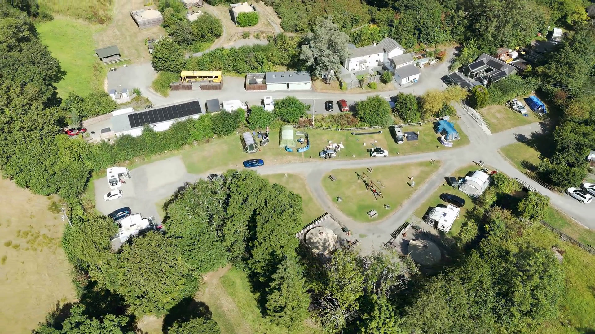 Looking straight down on the camping field in summer: tents, caravans and campervans on the pitches, the swings on the grass triangle, the yellow school bus by the lane at the top, and two bell tents on their decks at the bottom