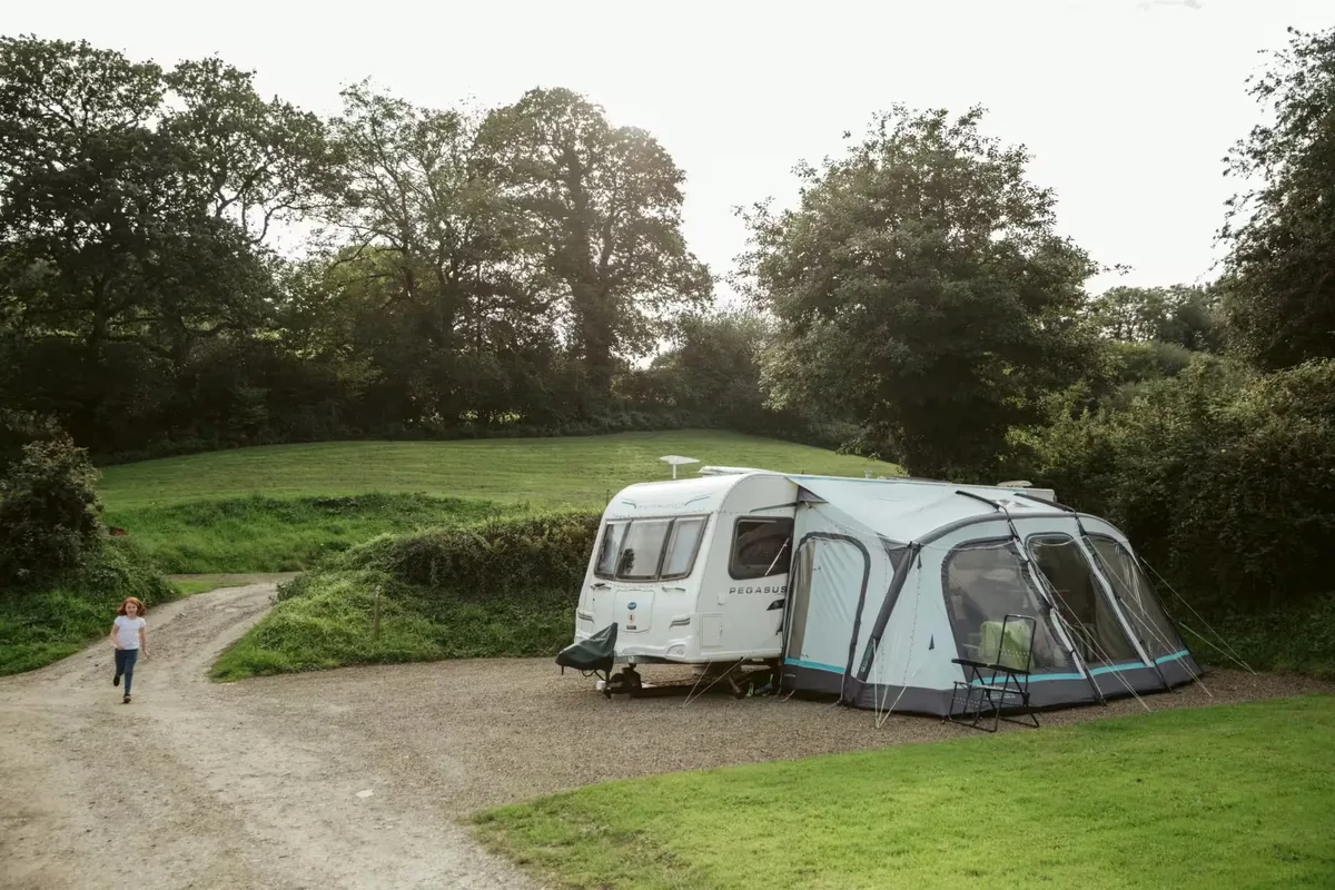 A touring caravan with its awning up on a gravel pitch at Willow Valley, the track curving past it and the grass bank behind